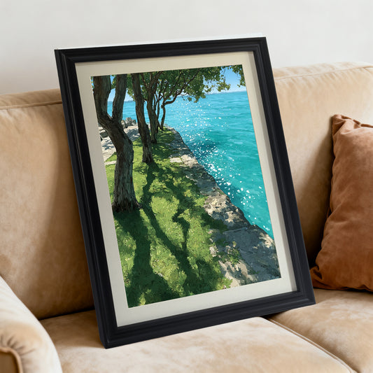A black-framed art print resting diagonally on a tan, plush sofa next to a brown pillow. The artwork features trees casting shadows on green grass by bright, sparkling blue water.