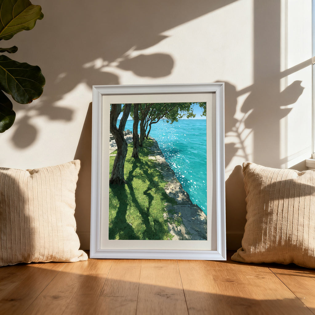 A white-framed art print leaning on a wooden floor between two cream-colored pillows. The print shows a sunny waterside scene with trees, grass, and bright blue water. Natural light and deep shadows define the scene.