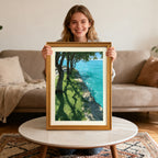 A smiling woman holding a vertical wood-framed art print above a round coffee table in a living room. The print features a vibrant waterside scene with trees, grass, and sparkling turquoise water.