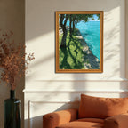 A wood-framed art print hanging on a cream-colored wall above an orange armchair. The print shows trees, grass with long shadows, and sparkling turquoise water. Sunlight streams in from the side.