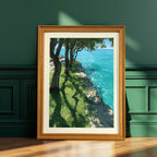 A vertical wood-framed art print leaning against a dark teal, paneled wall. The artwork depicts a sunny scene with trees casting long shadows on green grass next to bright turquoise water.