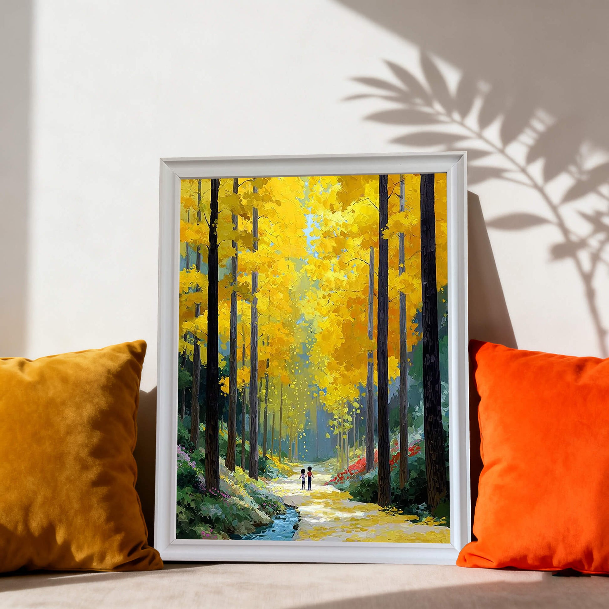 The forest painting in a white frame resting on a floor or low shelf between a yellow velvet pillow and an orange pillow.
