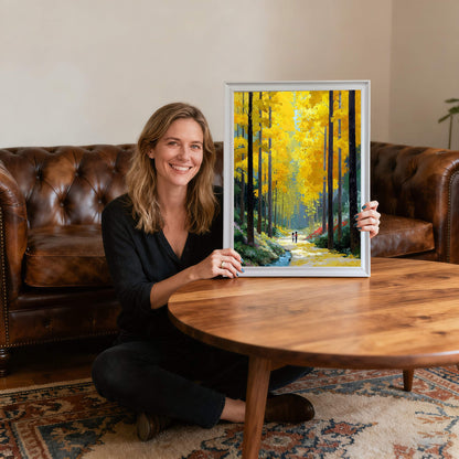 A woman sitting on a rug in a living room, smiling and holding the white-framed forest painting next to a round wooden coffee table.