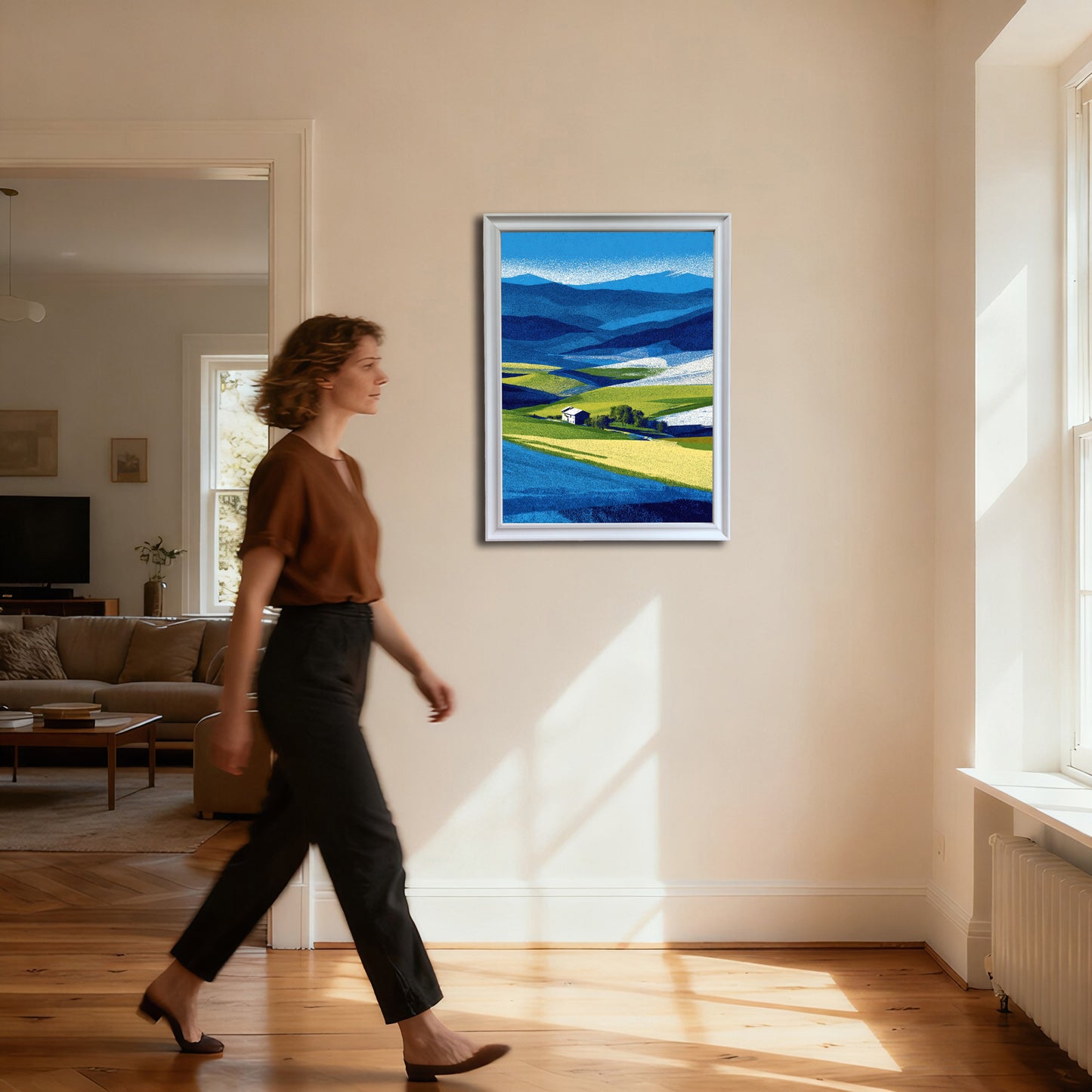 Full-body shot of a woman walking past a vertical abstract farm landscape print in a thin white frame, hanging on a cream wall in a brightly lit hallway.