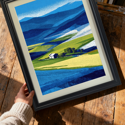Close-up shot of hands holding a framed abstract farm landscape print with bright blue and yellow fields, in a thick black wood frame, over a rustic wooden table.