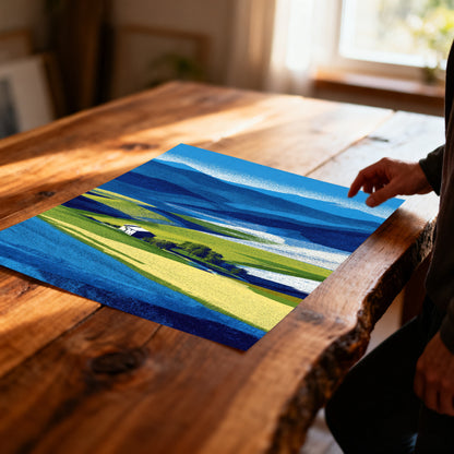 Overhead shot of a hand reaching toward an unframed abstract print of a hilly farm landscape with deep blue mountains and bright yellow-green fields, resting on a rustic wood table.