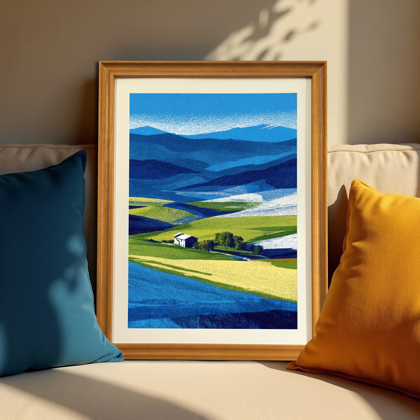 Vertical abstract farm landscape print in a natural brown wood frame, resting on a neutral-colored couch between a dark blue and a bright yellow throw pillow.