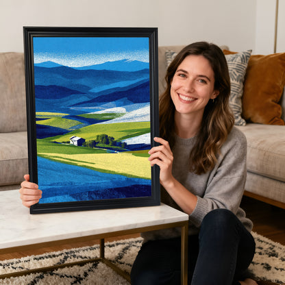 A smiling woman sitting on the floor holding a framed abstract farm landscape print in a thick black wood frame, with a white coffee table and sofa in the background.