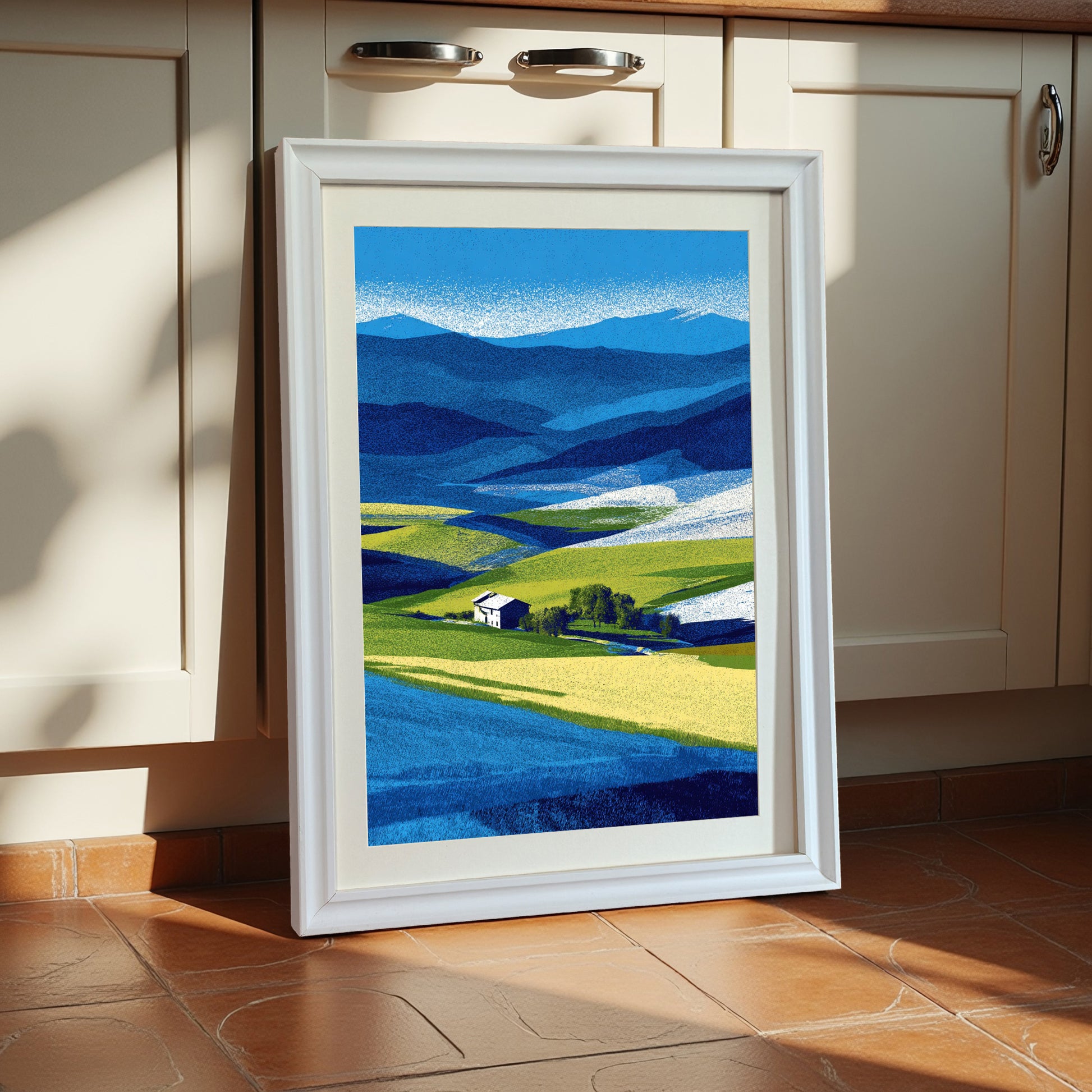 Vertical abstract farm landscape print with blue mountains, bright yellow fields, and a white farmhouse, framed in a thick white wooden frame, leaning against cream kitchen cabinets.