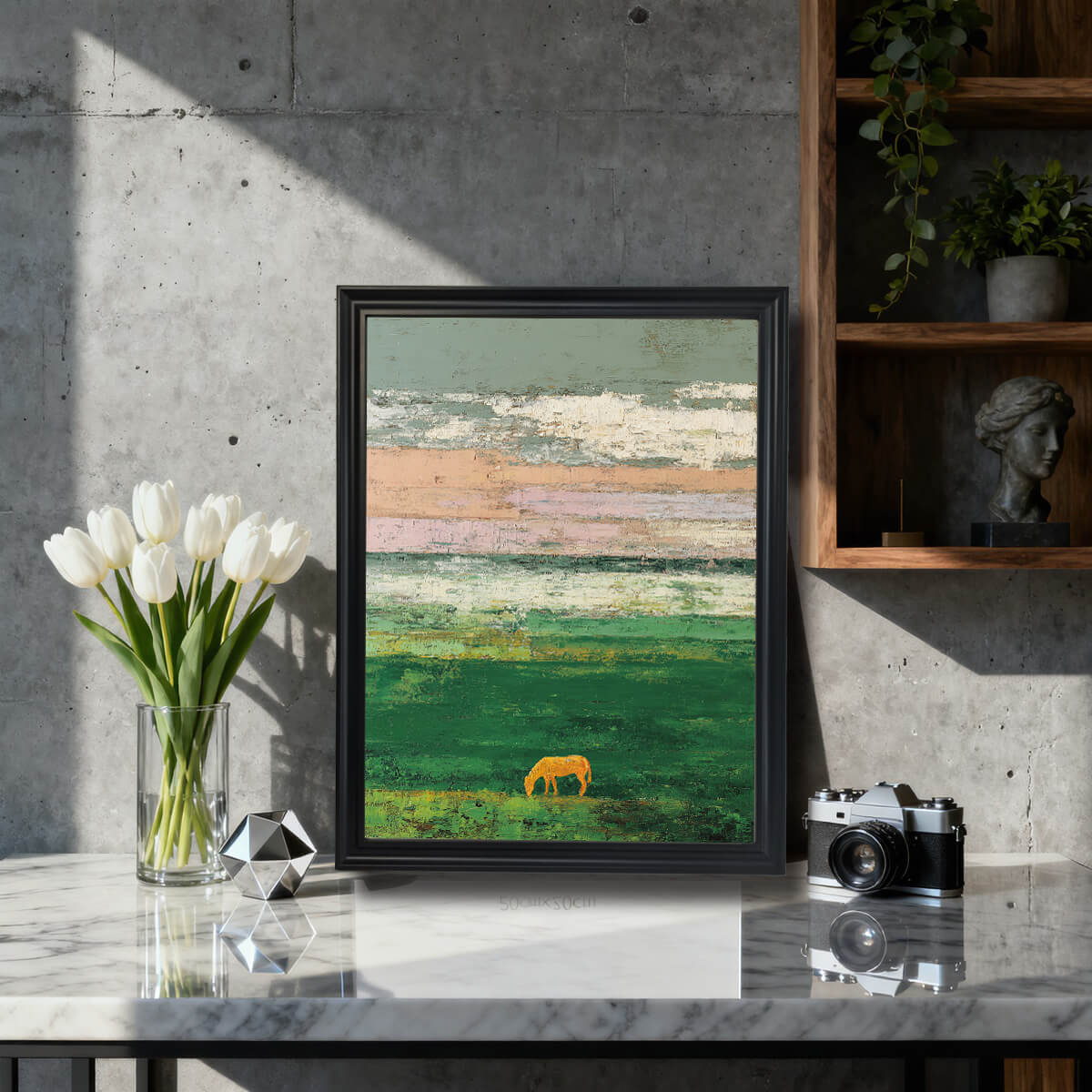 A framed art print of an abstract landscape with textured horizontal bands of pale pink, grey, and white, over a vibrant green field where a golden horse silhouette grazes. The print is in a black frame, resting on a marble-top table next to white tulips in a modern, concrete-walled room.