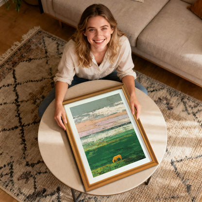 A smiling woman sits on the floor holding a framed art print of an abstract green field with a grazing golden horse silhouette and a textured, colorful sky. The frame is natural wood and rests on a round coffee table.