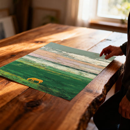 A close-up, angled view of an unframed art print lying on a rustic wooden table. The print shows an abstract green field with a golden horse silhouette and textured sky bands. A person's hand is visible on the right.