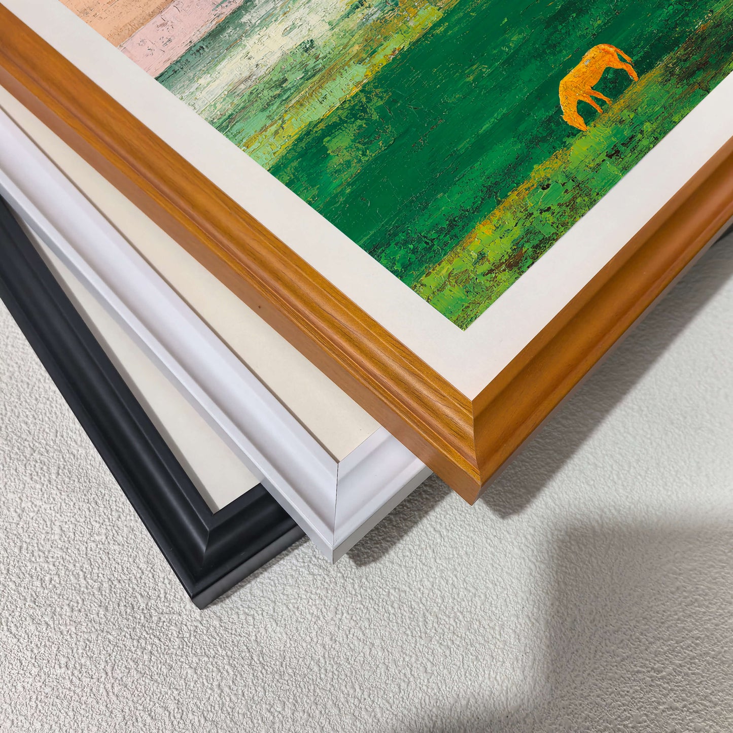 A stack of wood picture frame corners showing different color options: natural wood, white, cream/off-white, and black. A corner of the abstract art print, showing the textured green field and golden horse, is visible on top.