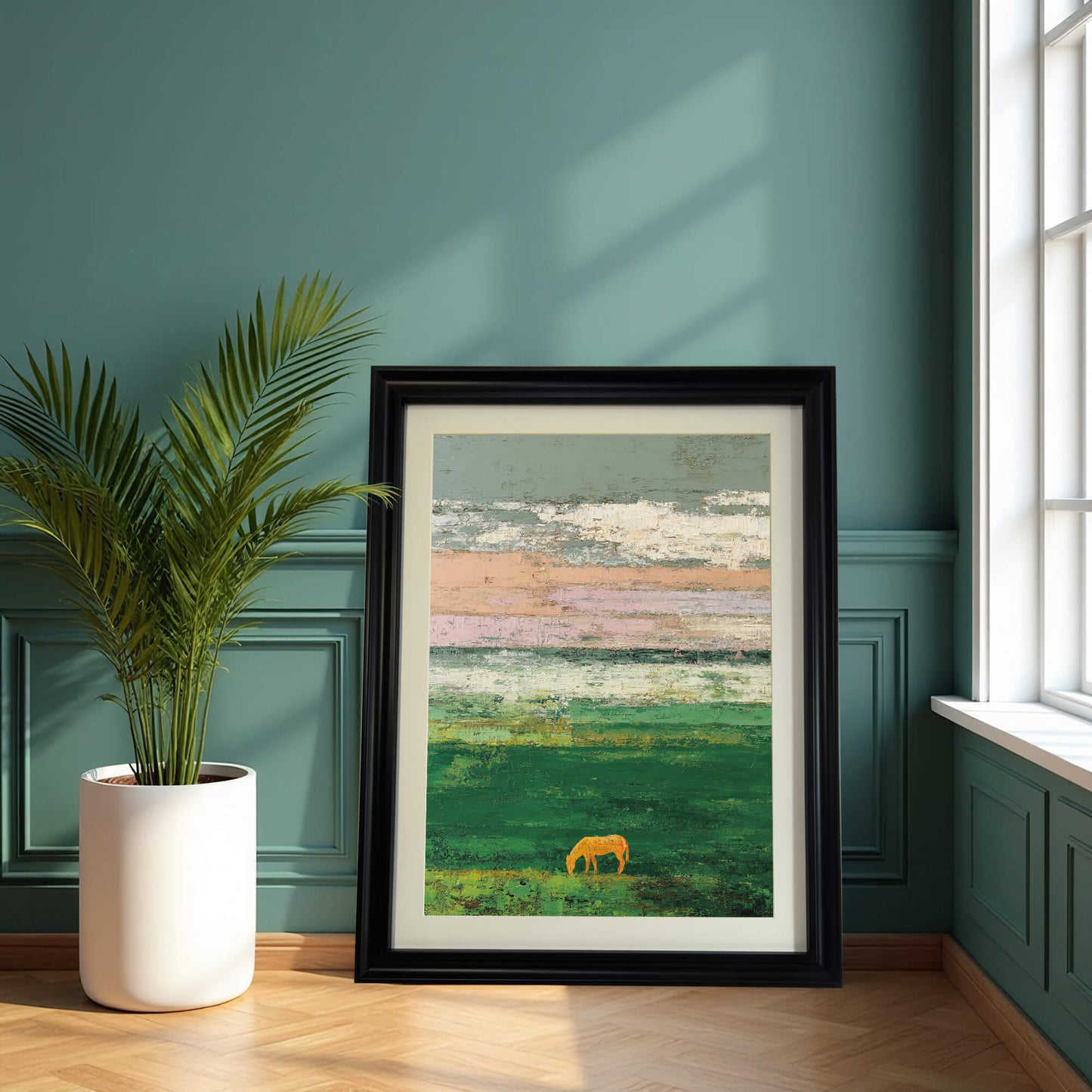 A framed art print of an abstract green field and sky with a golden horse silhouette, resting on a wooden floor against a dark, paneled green wall, beside a tall potted palm plant. The frame is black.