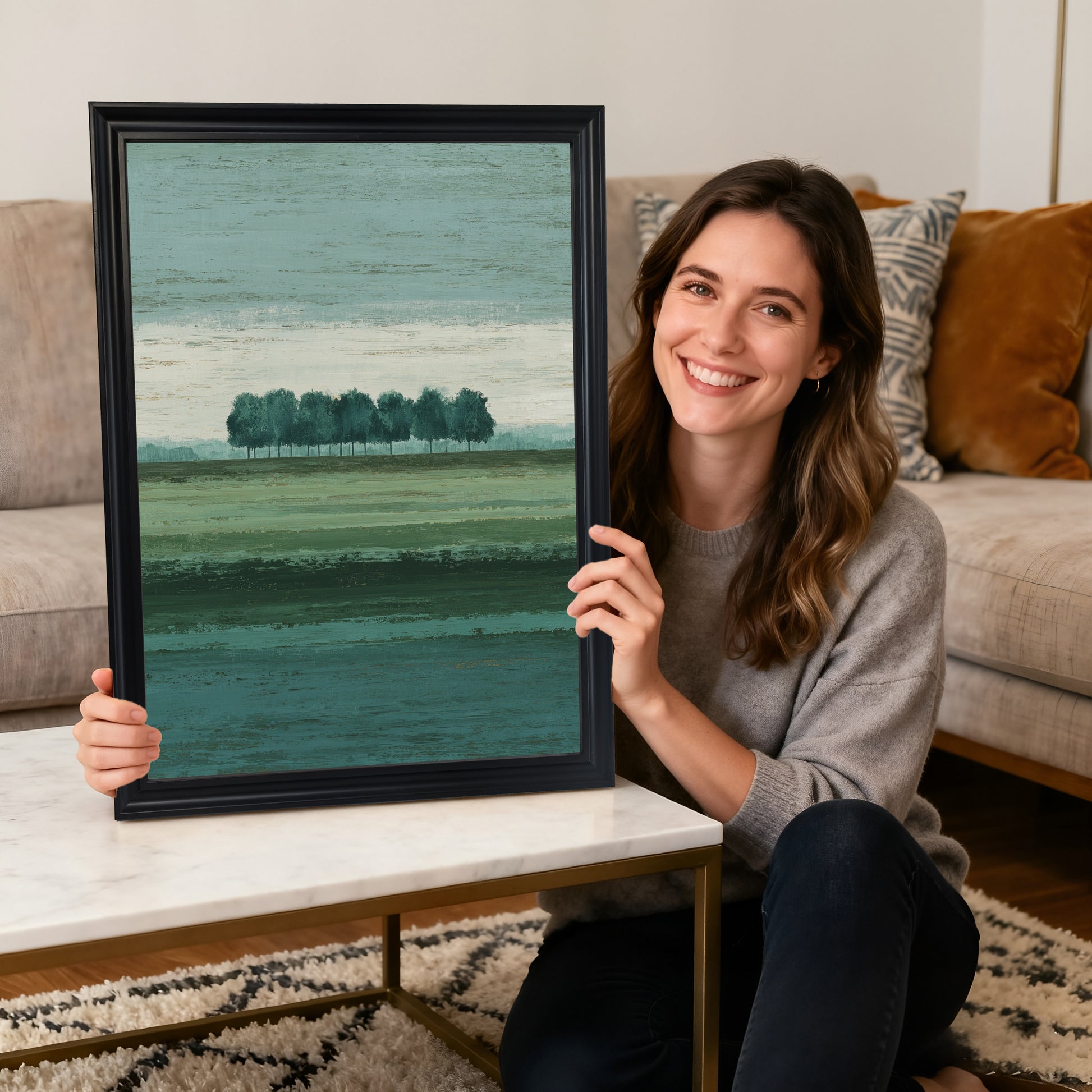 A smiling woman sitting on the floor holding a vertical abstract landscape art print with the row of trees motif, framed in a thick black wood frame, with a white coffee table and sofa in the background.