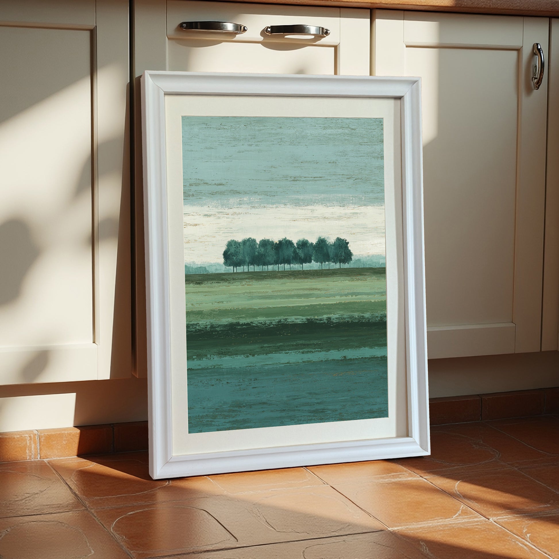 Vertical abstract landscape art print showing a row of trees over banded green fields, framed in a thick white wooden frame with a mat, leaning against cream kitchen cabinets.