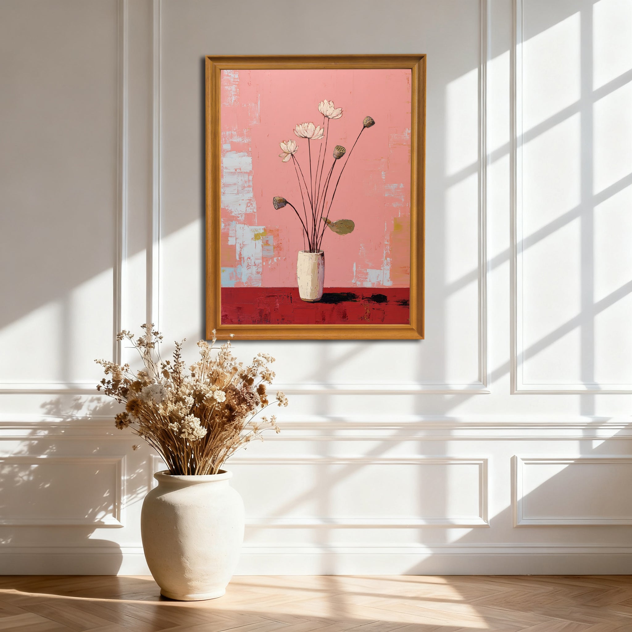 A vertical, gold-framed painting of white lotus flowers on a pink and red background, hanging on a white wainscoted wall. A large, off-white ceramic vase with dried flowers sits on the wooden floor below it, with strong sunlight creating window shadows.