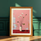 A vertical, framed painting of white lotus flowers in a white vase against a pink and red background. The painting is in a light brown wood frame and is leaning on a hardwood floor against a dark green paneled wall with strong window light.