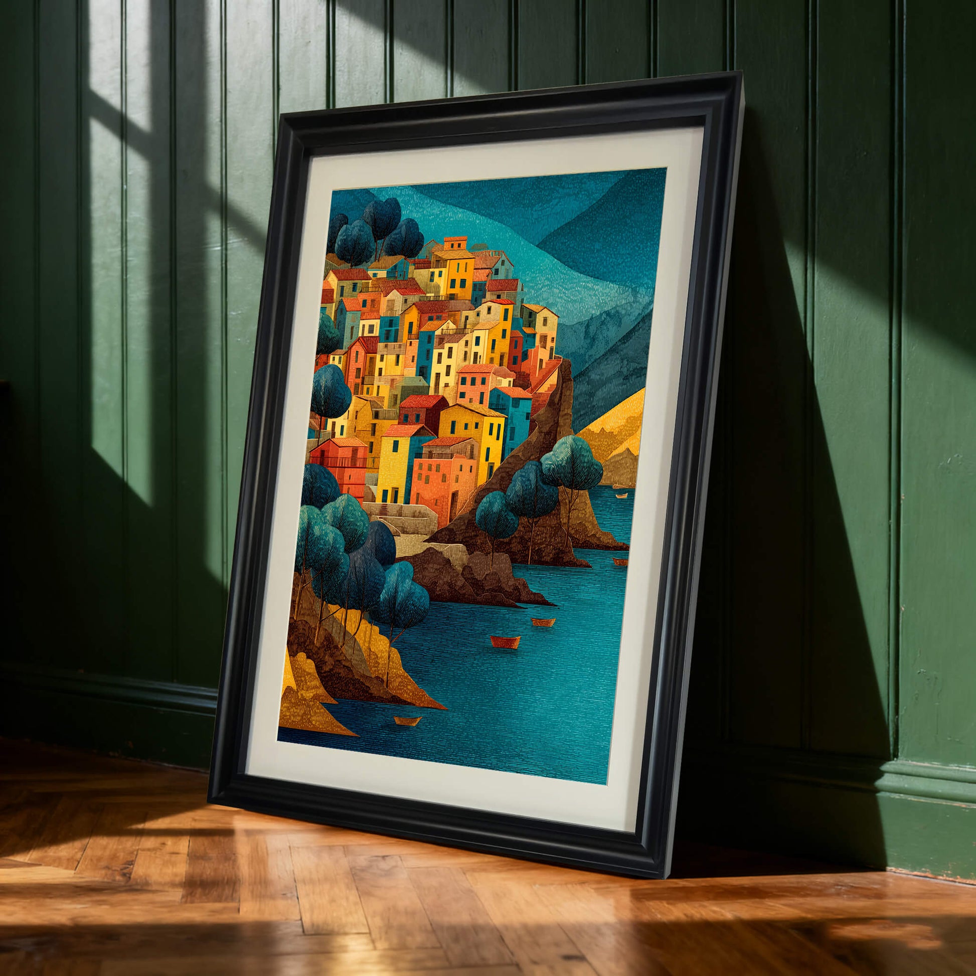Art print of a colorful cliffside village in a black frame, leaning on a wooden floor against a dark green paneled wall.