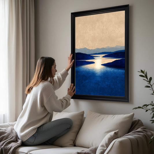 A woman in a white sweater adjusting a large framed art print of the blue lake landscape in a black frame, hanging above a white sofa.