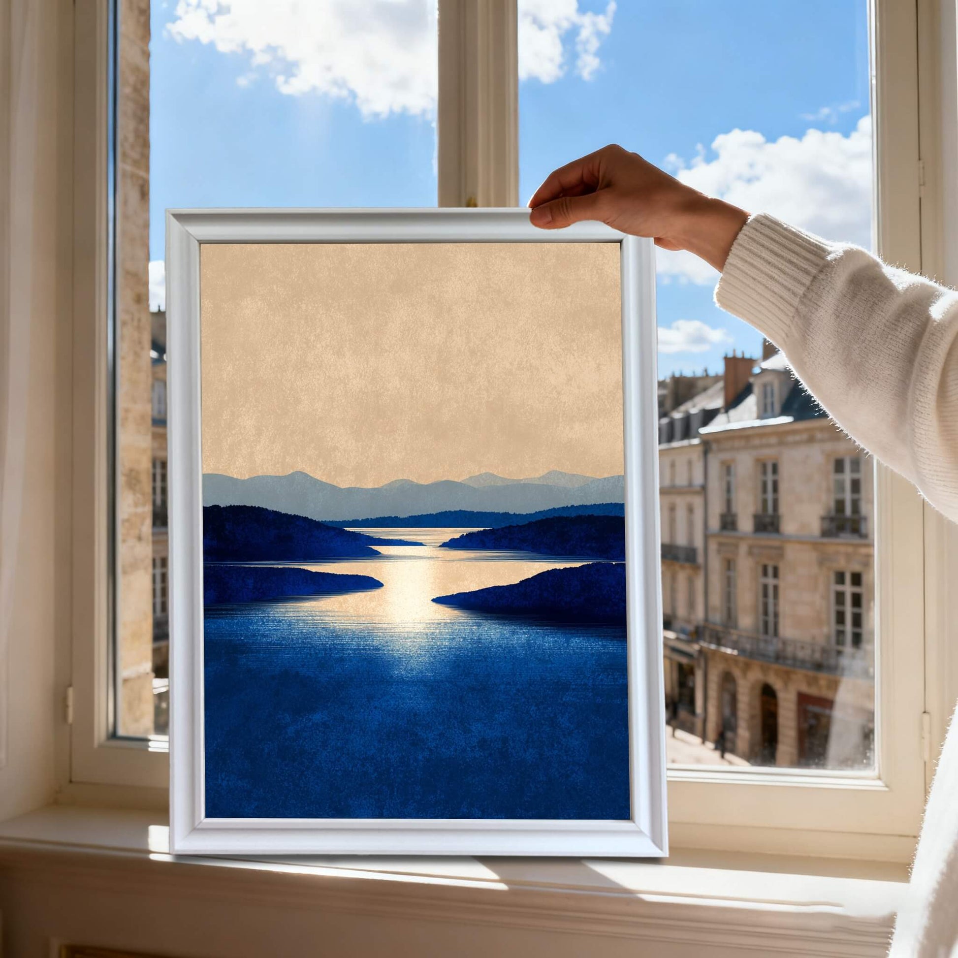 Hands holding the framed art print of the blue lake landscape in a white frame, silhouetted against a bright sunny window overlooking a city street.