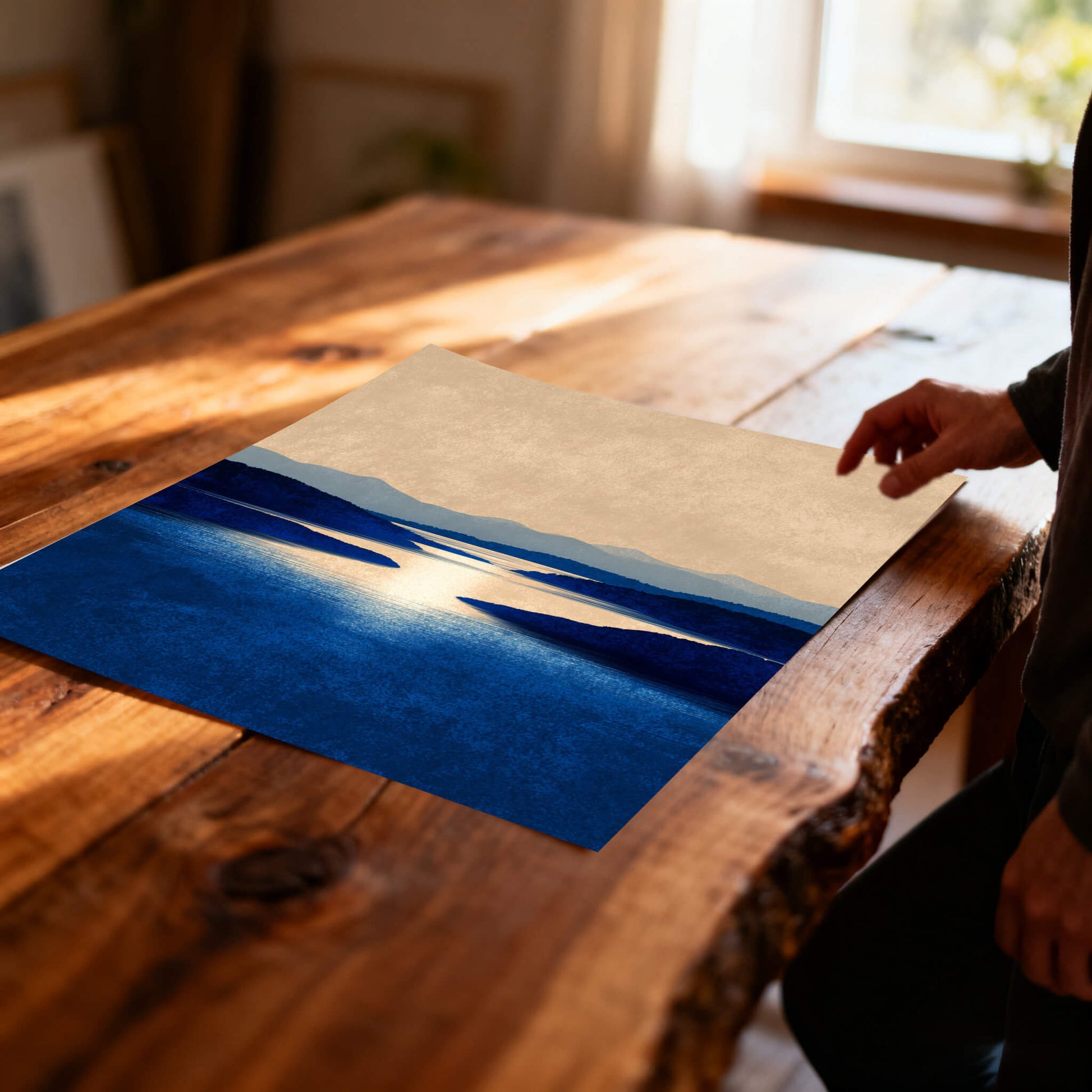 Unframed art print of the blue lake and hills landscape print lying on a rustic, live-edge wooden table in soft sunlight.