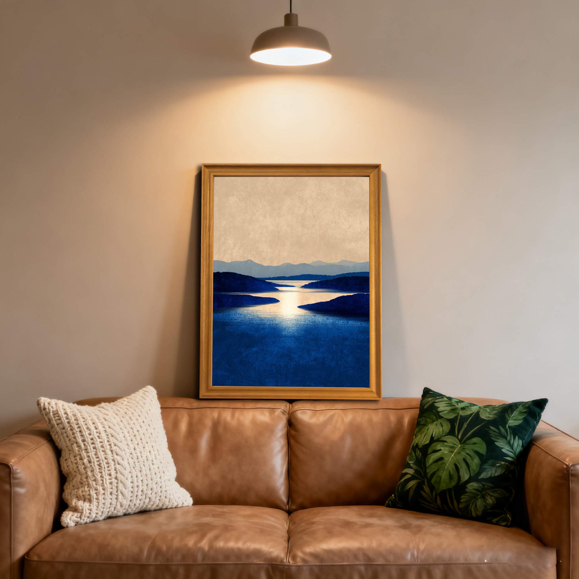 Framed art print of a serene blue lake and hills in a light wood (natural) frame, hanging over a brown leather couch with textured and tropical print pillows.