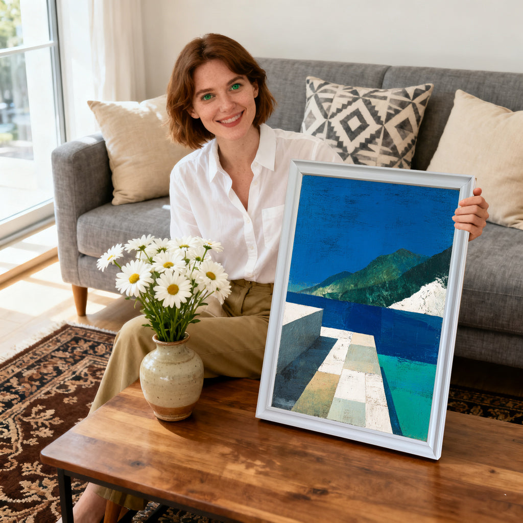 Smiling woman sitting on a couch, holding a framed abstract landscape art print with blue, green, and white tones. The frame is white. A vase of daisies is on the coffee table.