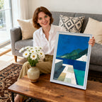 Smiling woman sitting on a couch, holding a framed abstract landscape art print with blue, green, and white tones. The frame is white. A vase of daisies is on the coffee table.
