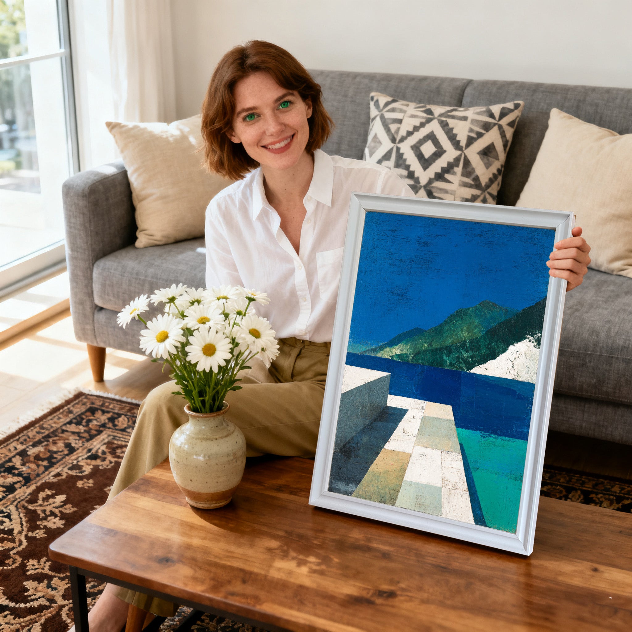Smiling woman sitting on a couch, holding a framed abstract landscape art print with blue, green, and white tones. The frame is white. A vase of daisies is on the coffee table.