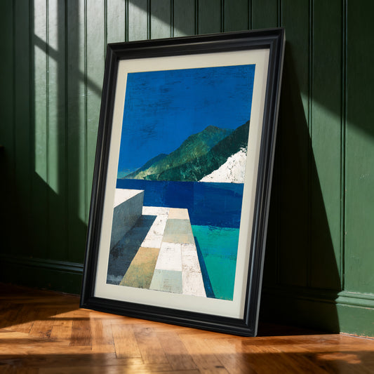 Framed abstract landscape art print with a black frame leaning against a dark green paneled wall. Natural light casts dramatic shadows on the wall and wooden floor.