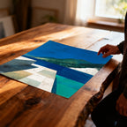 Abstract landscape art print featuring blue water and green mountains, resting unframed on a natural wood plank table with a person's hand near the edge.