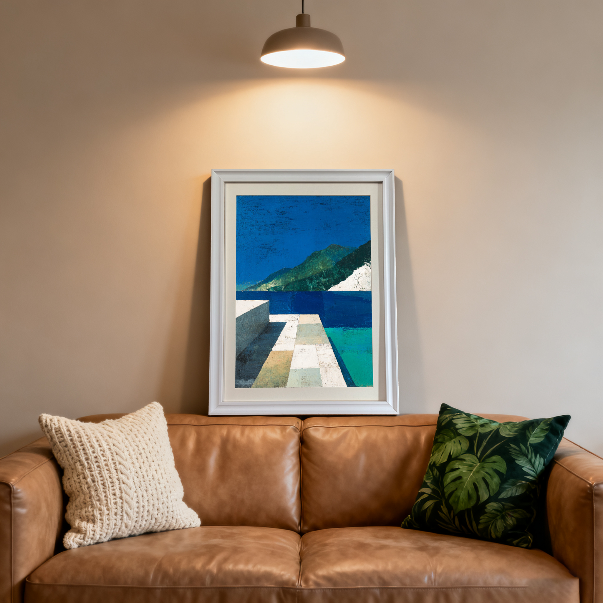 White-framed abstract landscape art print centered above a brown leather couch with two decorative pillows. The print is highlighted by a ceiling spotlight.