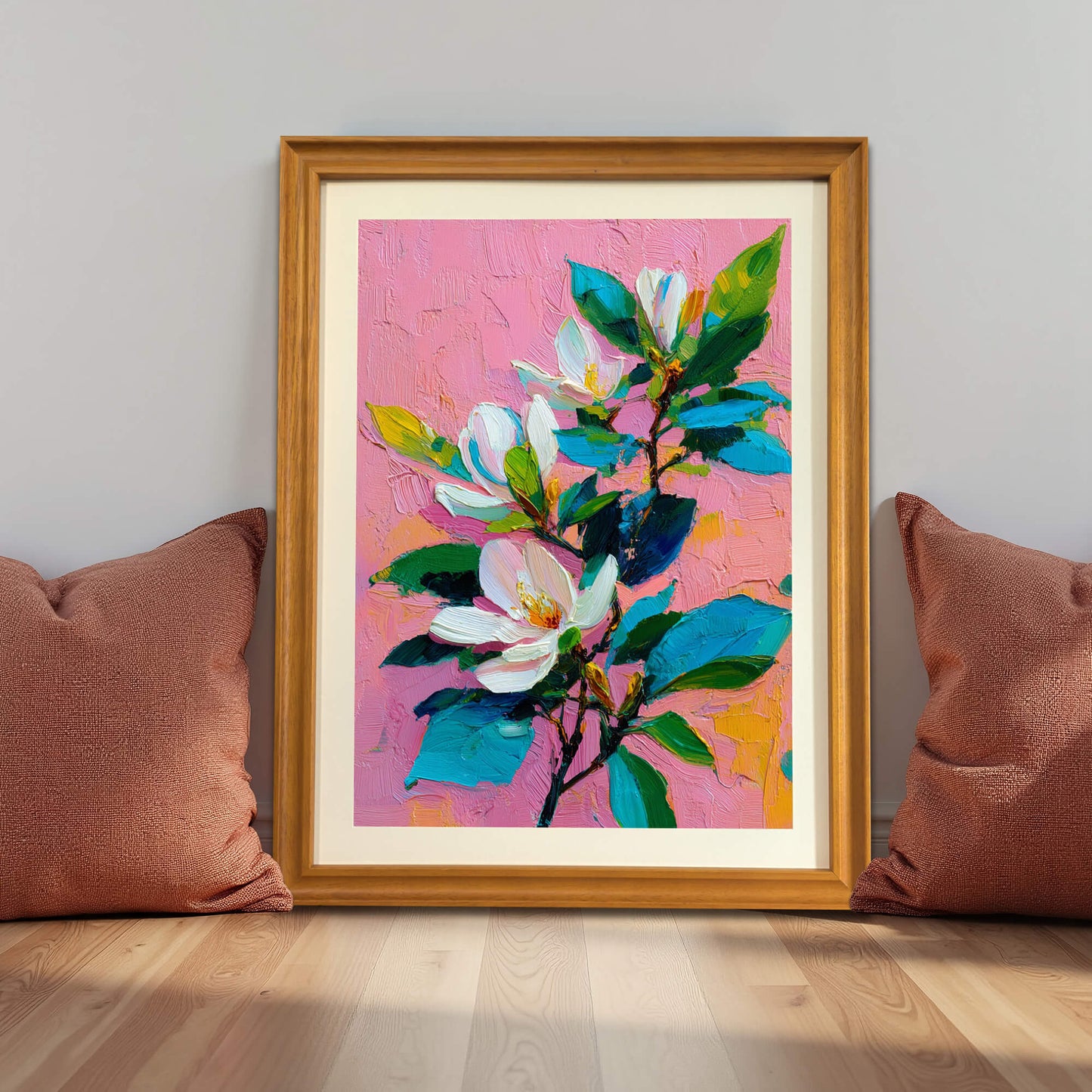 Wood-framed floral art print of white magnolias on pink, placed on a floor between two brown pillows.