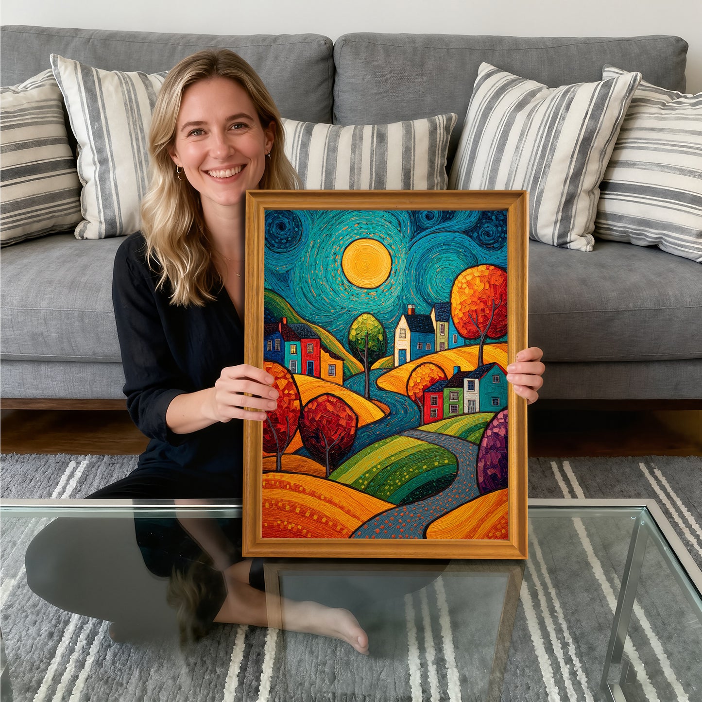 A smiling woman sitting on the floor in front of a gray couch, holding the vibrant village art print framed in gold-toned wood, showcasing the artwork.