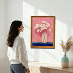 A woman in a white sweater looking at an oil painting on a white wall. The painting features white flowers in a clear bottle, set against a textured, vibrant pink background with a dark blue foreground, framed in a gold/natural wood frame.