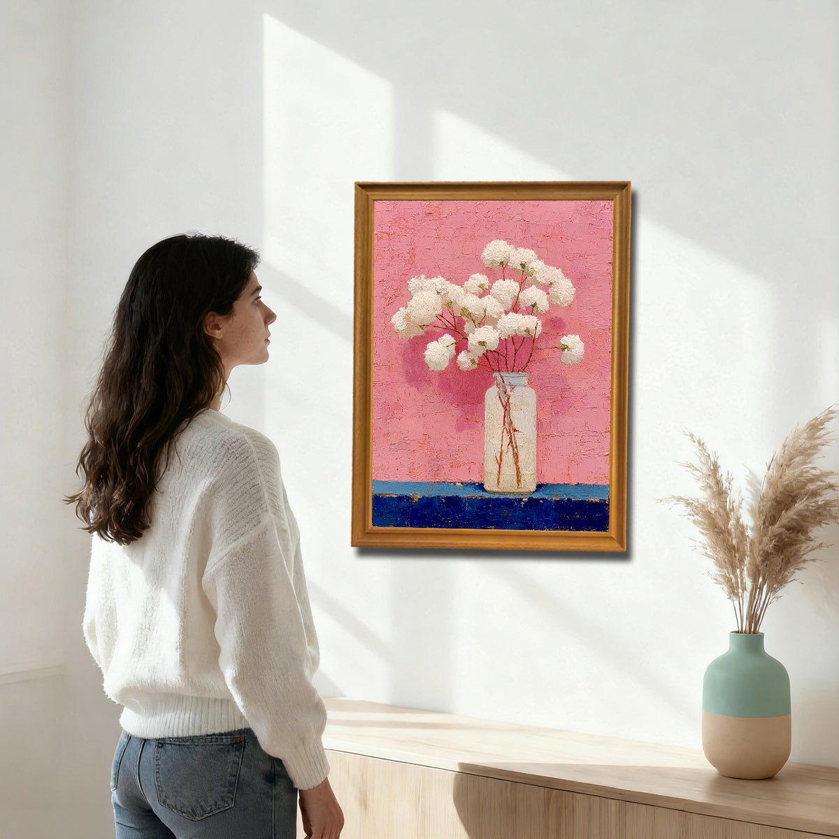 A woman in a white sweater looking at an oil painting on a white wall. The painting features white flowers in a clear bottle, set against a textured, vibrant pink background with a dark blue foreground, framed in a gold/natural wood frame.