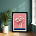 The framed art print, in a thick black frame, is leaning against a dark teal wall with wainscoting details, next to a large potted plant and a bright window. The painting depicts white flowers on a pink background.