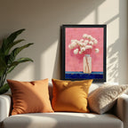 The art print, framed in a sleek black frame, is displayed in a modern living room setting, resting on a console behind a white sofa with orange and brown pillows. Sunlight casts shadows across the wall.