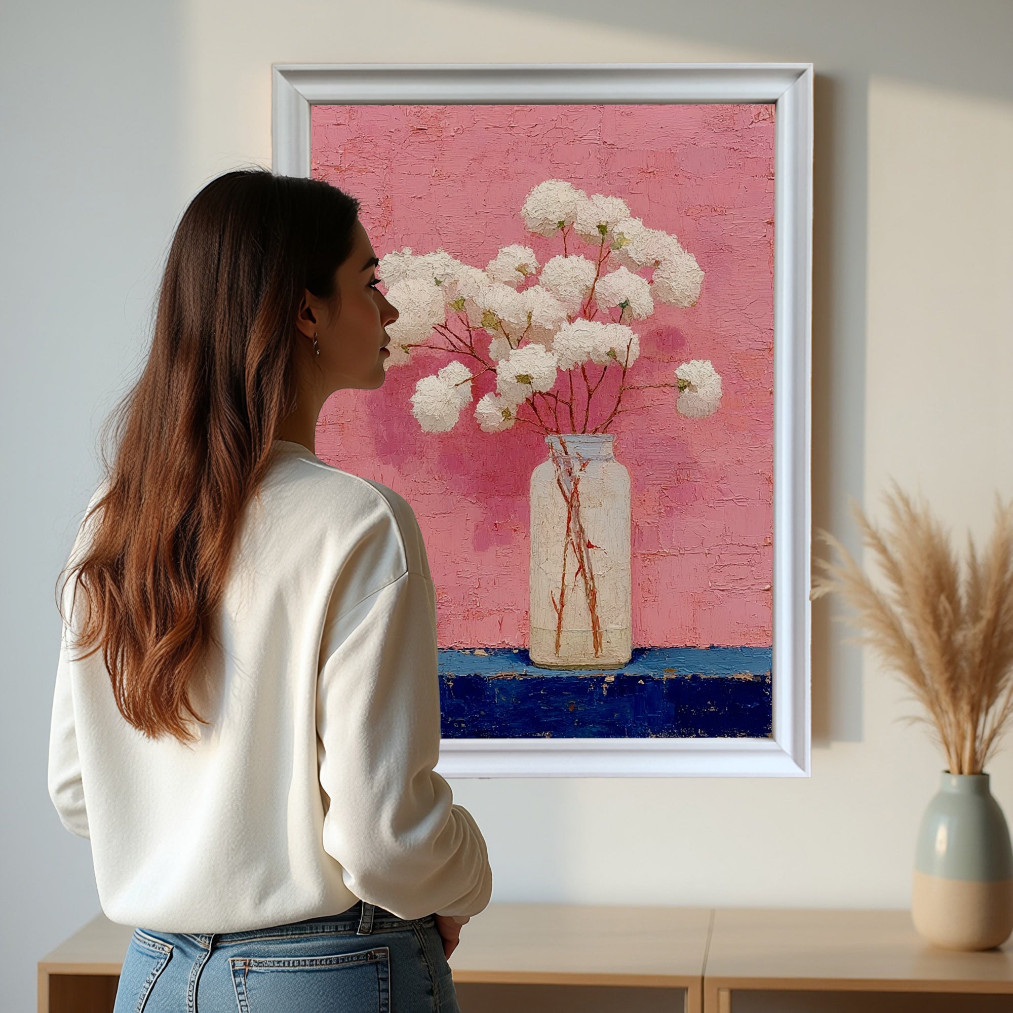 A woman in a white sweatshirt looking intently at the framed art print. The painting features white flowers in a bottle against a textured pink background and a dark blue base, set in a clean, contemporary white frame.
