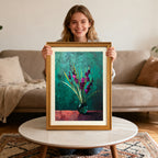 Smiling woman holding up a framed art print of purple flowers in a vase with a gold-colored wood frame, sitting behind a round marble coffee table.