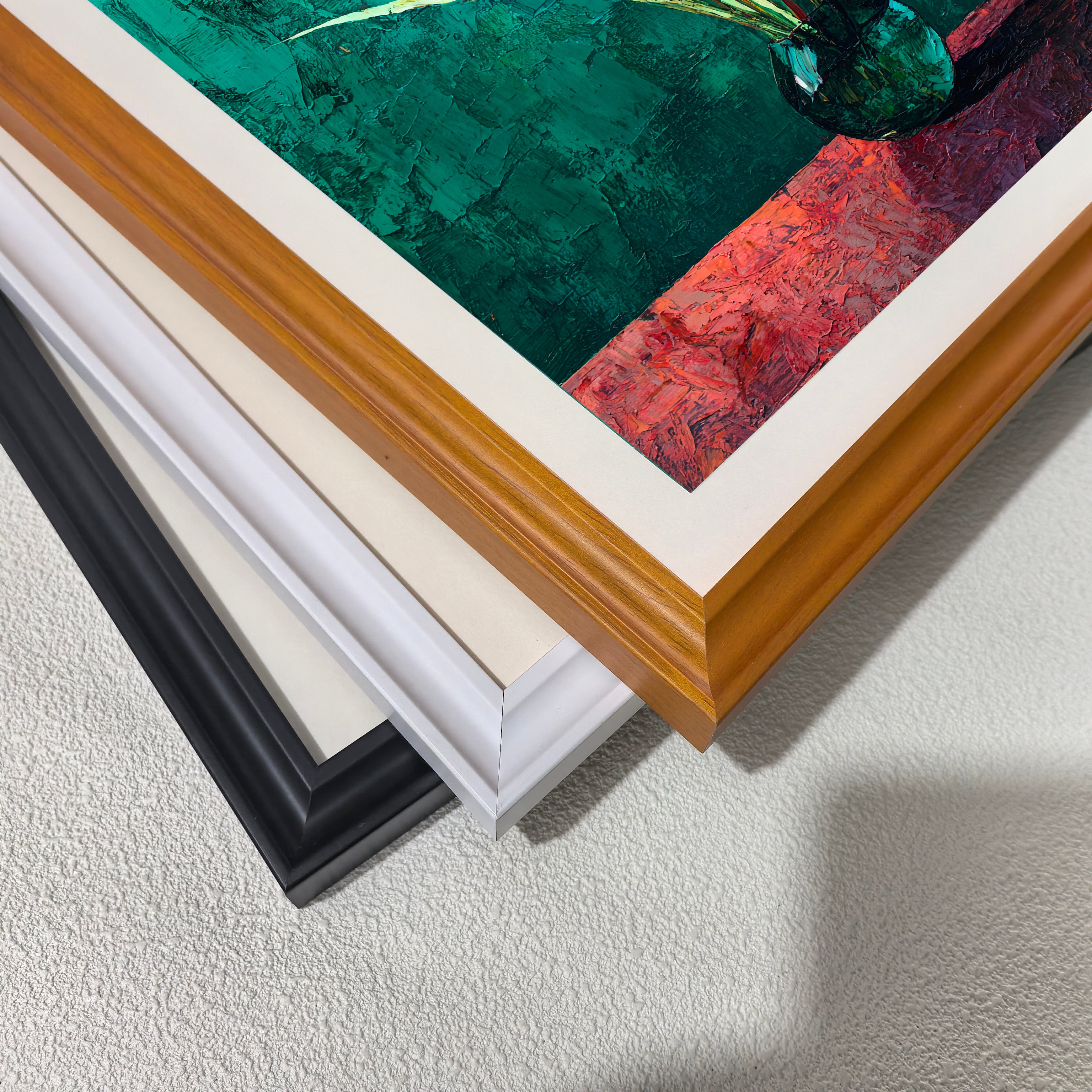 Close-up of three stacked wood picture frame corners in black, white, and natural brown, showing the art print inside which features dark green and red colors.
