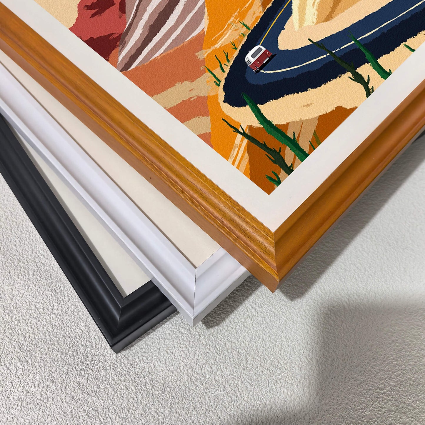 A corner close-up showing the detail of the desert road artwork and three stacked frame options: warm wood, white, and black, demonstrating the available wood frame styles.