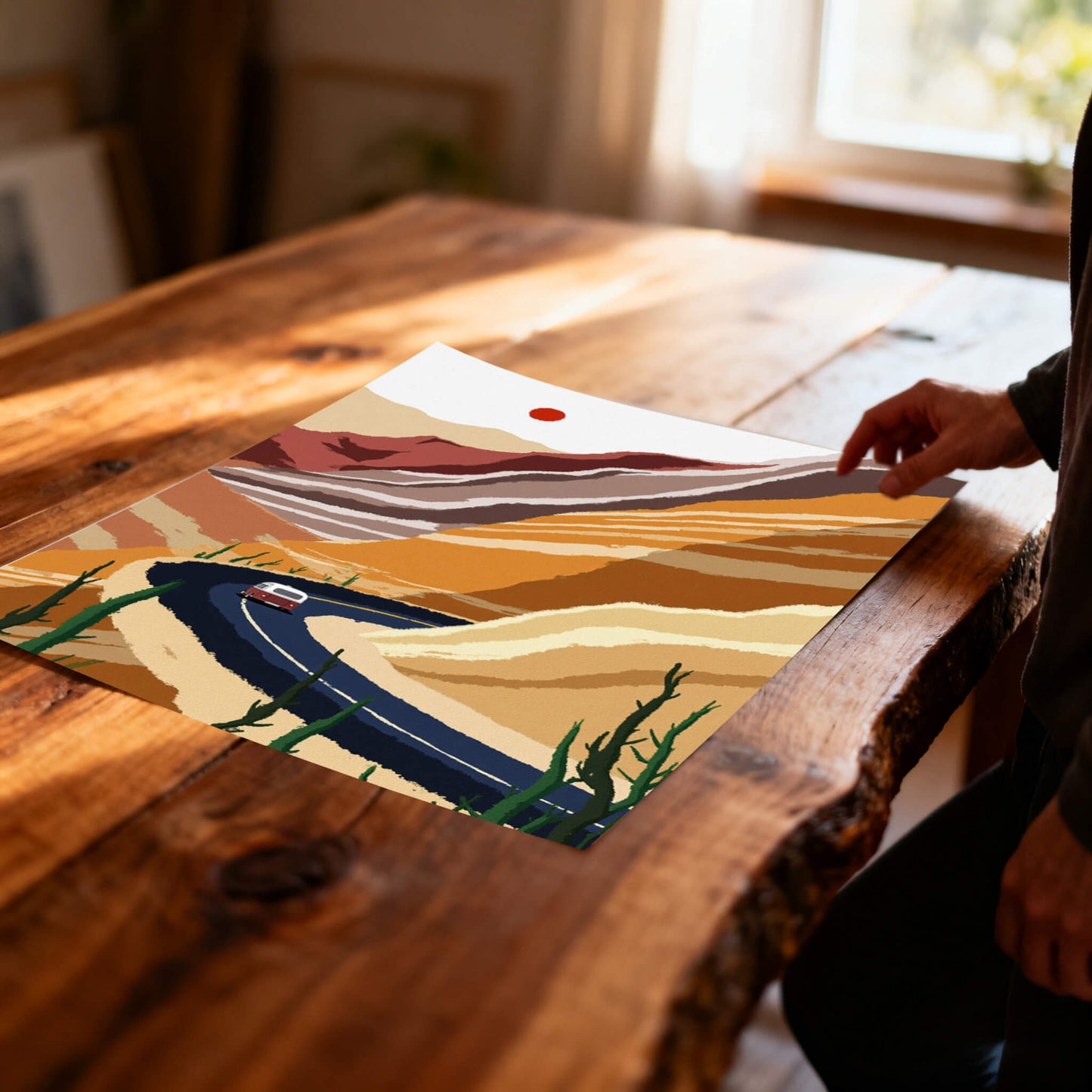 A close-up of the unframed desert landscape print lying flat on a rustic wooden table, showing a person's hand touching the corner near a sun-drenched window.