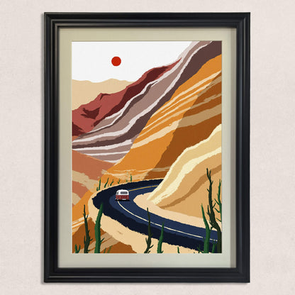 A close-up of the framed desert landscape art print, featuring a red van on a dark winding road against striated yellow, brown, and white hills, framed in a detailed black wood frame with a mat.