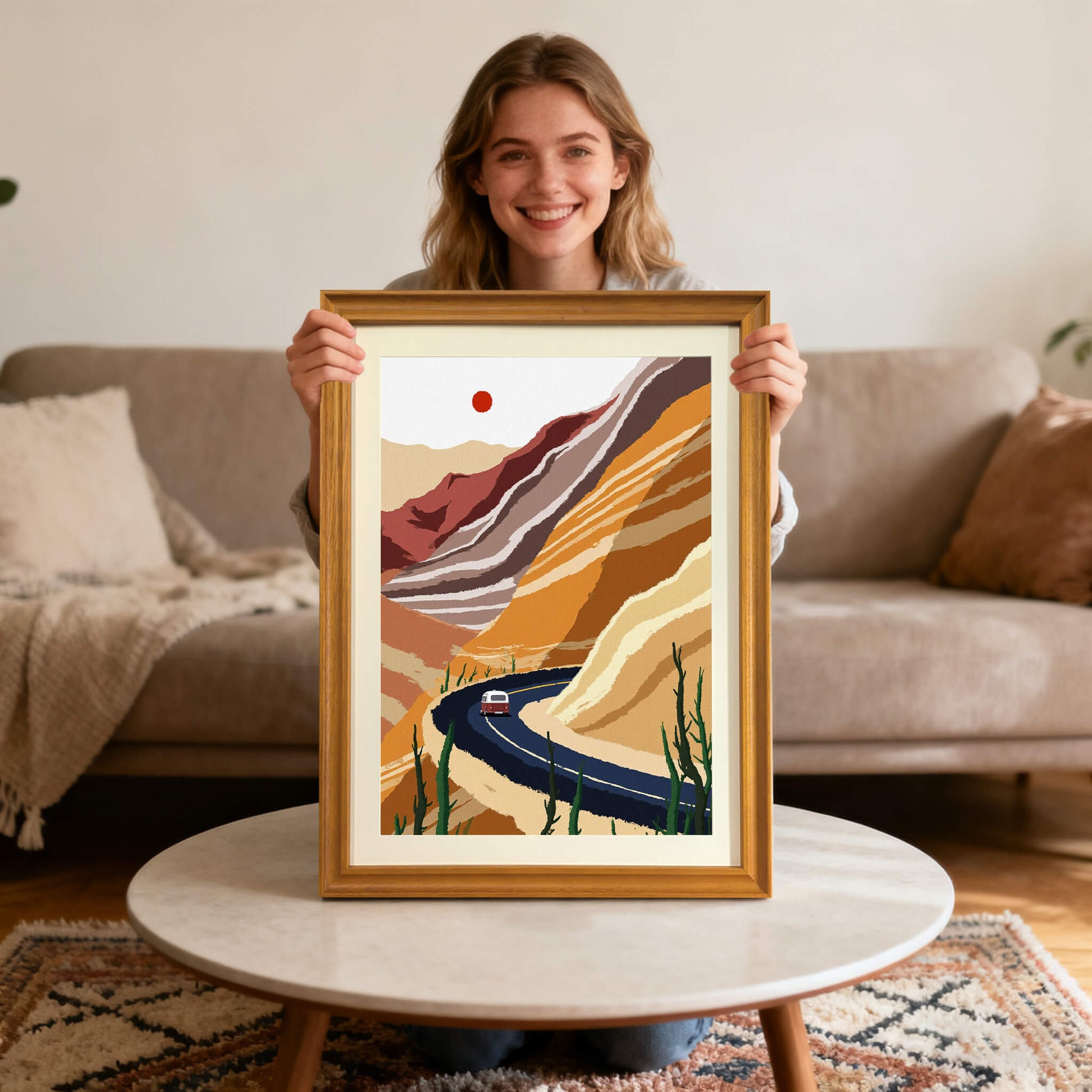 A smiling man holding the desert landscape art print, framed in warm wood, while sitting on a wooden bench in front of a bright, sunlit window overlooking the ocean.