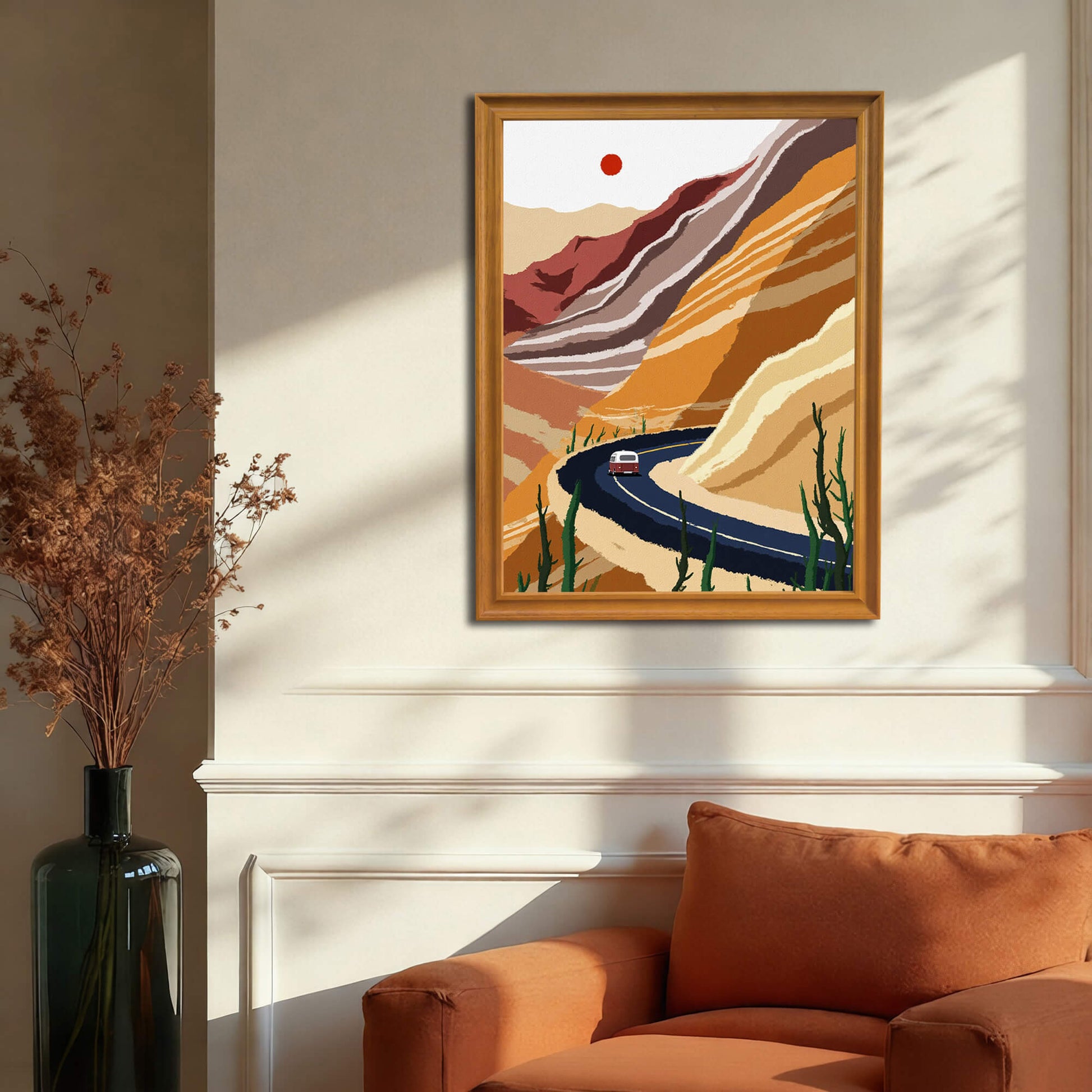 The desert road art print, framed in warm wood, hanging on a cream wainscoting wall above a rust-orange armchair. A dark vase with dried flora stands next to the chair.