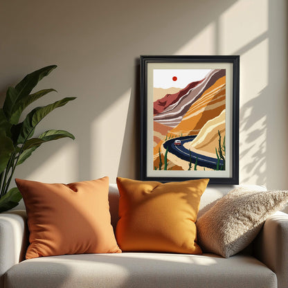 The desert landscape art print, framed in black wood, standing on a white couch between two orange pillows, next to a large potted plant. Sunlight streams in, casting sharp shadows.
