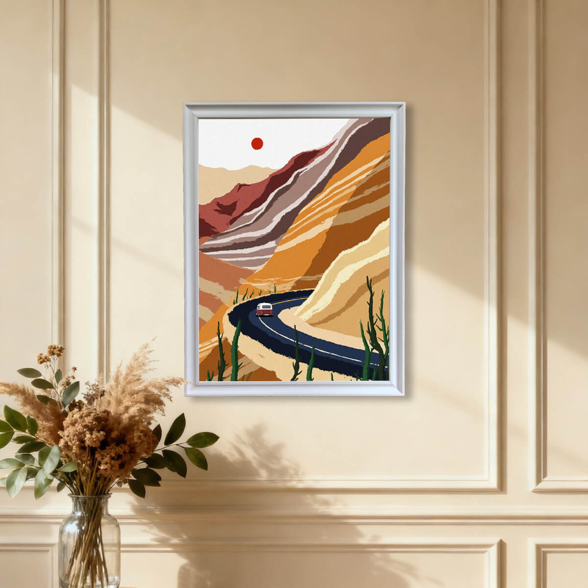 The modern desert landscape art print featuring a winding road and layered rock formations, framed in white, hanging on a warm beige wall with wainscoting details, above a vase of dried pampas grass.