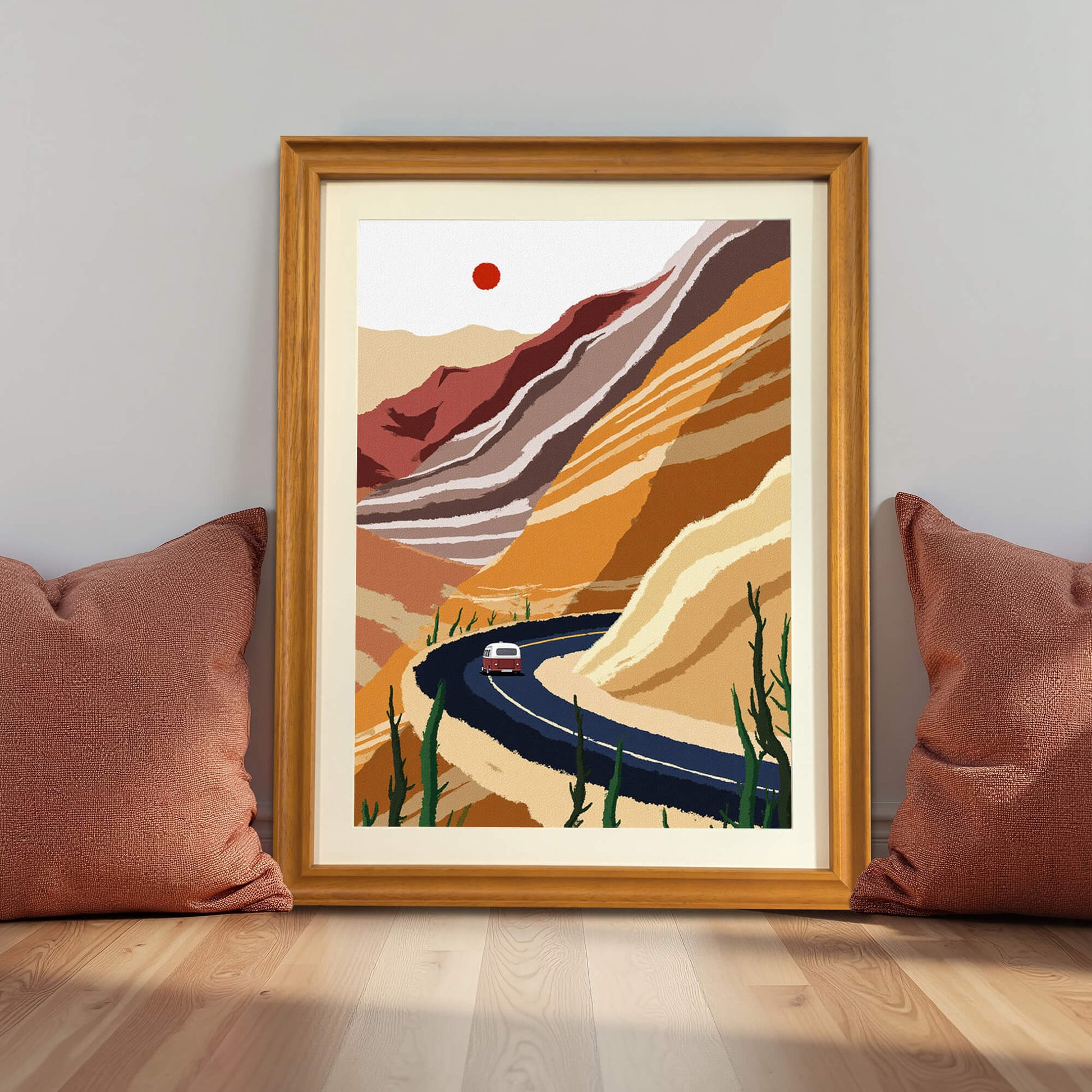 The desert landscape art print in a warm wooden frame, leaning on a wooden floor against a grey wall, flanked by two large, rust-colored throw pillows.